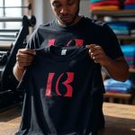 A person stands in a bright workshop, holding up a black T-shirt with a bold red logo on the chest. Another black T-shirt with the same logo lies folded on a wooden table in the foreground. Behind them, a printing press and shelves stacked with colorful folded shirts create a busy, creative atmosphere. Large windows let in natural light, illuminating the workspace.